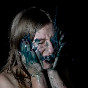 Eine junge Frau mit blonderem Haar, deren Gesicht von blauer Flüssigkeit bedeckt ist – verzerrt vor Emotion, Symbol für visuelles Storytelling und kreative Gestaltung.